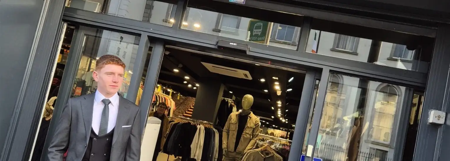 A man in a suit stands outside a clothes shop with glass doors. Inside, rails of clothes and a mannequin are visible. The shop is on a city street with buildings reflected in the windows.