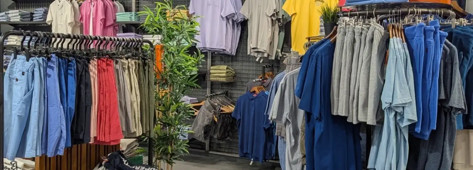 Clothing shop display with various rails of shirts and trousers in shades of blue, grey, pink, and beige. Some shirts are folded on shelves; a potted plant stands in the centre.