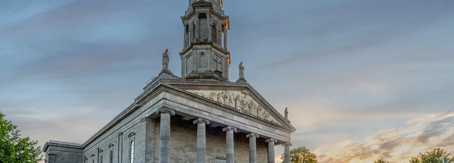 A historic stone building with tall columns, ornate statues on the roof, and a tall spire, set against a sunset sky with soft clouds and trees on either side.