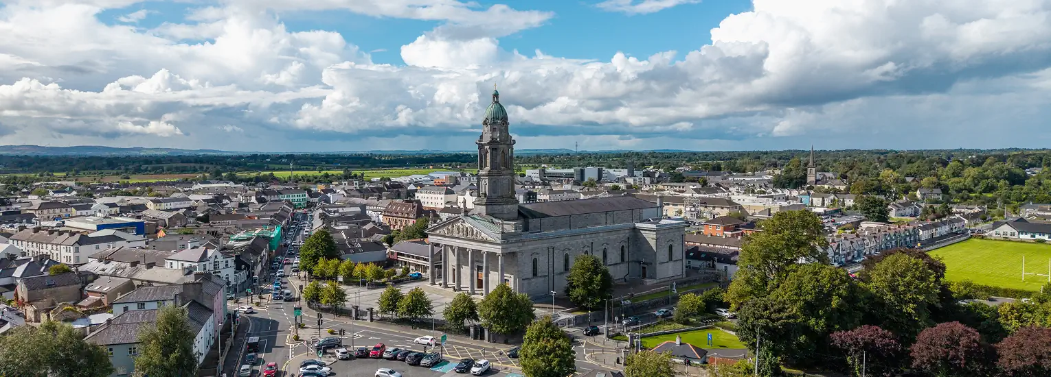 Aerial view of a large stone church with a tall clock tower surrounded by trees and buildings in a small town, with roads, cars, and green fields in the background under a partly cloudy sky.