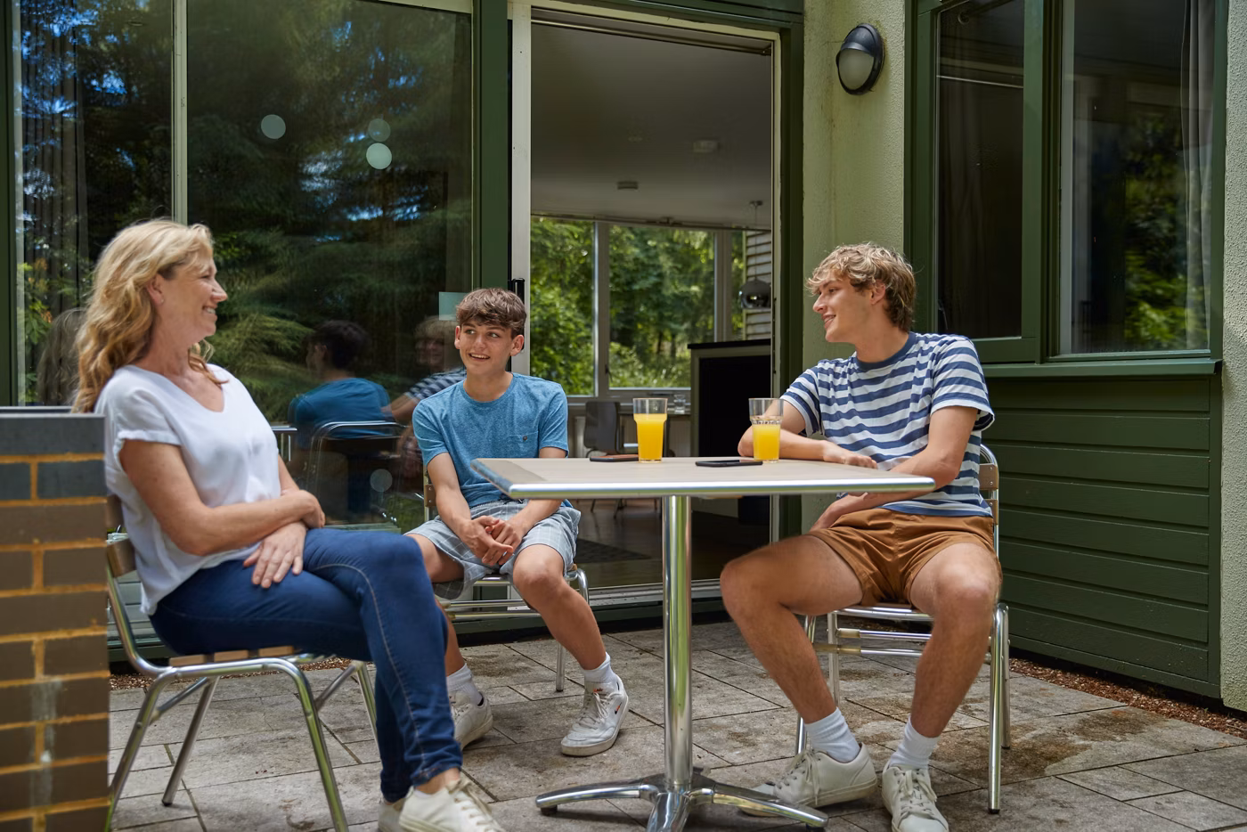 Three people sit outside at a round table with glasses of orange juice, smiling and talking on a sunny patio. There is greenery reflected in the glass doors and windows behind them.