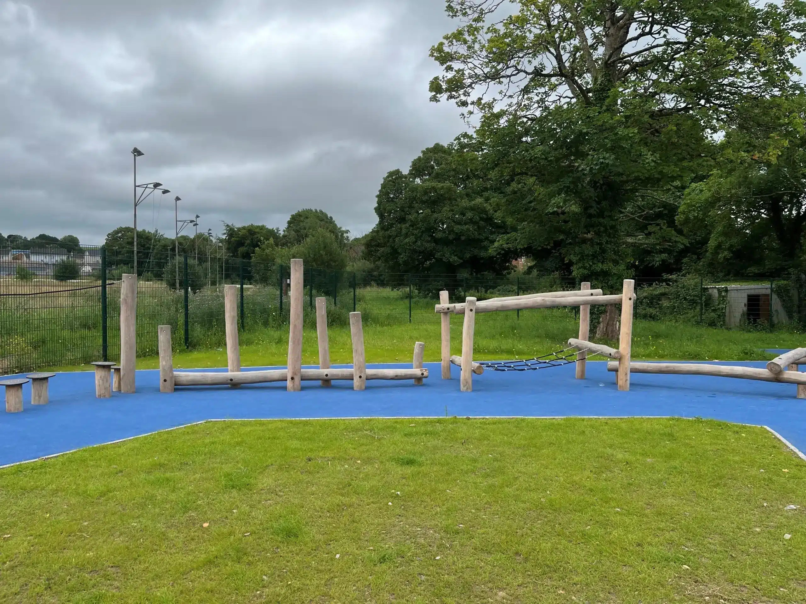 A wooden playground assault course with logs and chains sits on a blue rubber surface, surrounded by grass and trees under a cloudy sky.