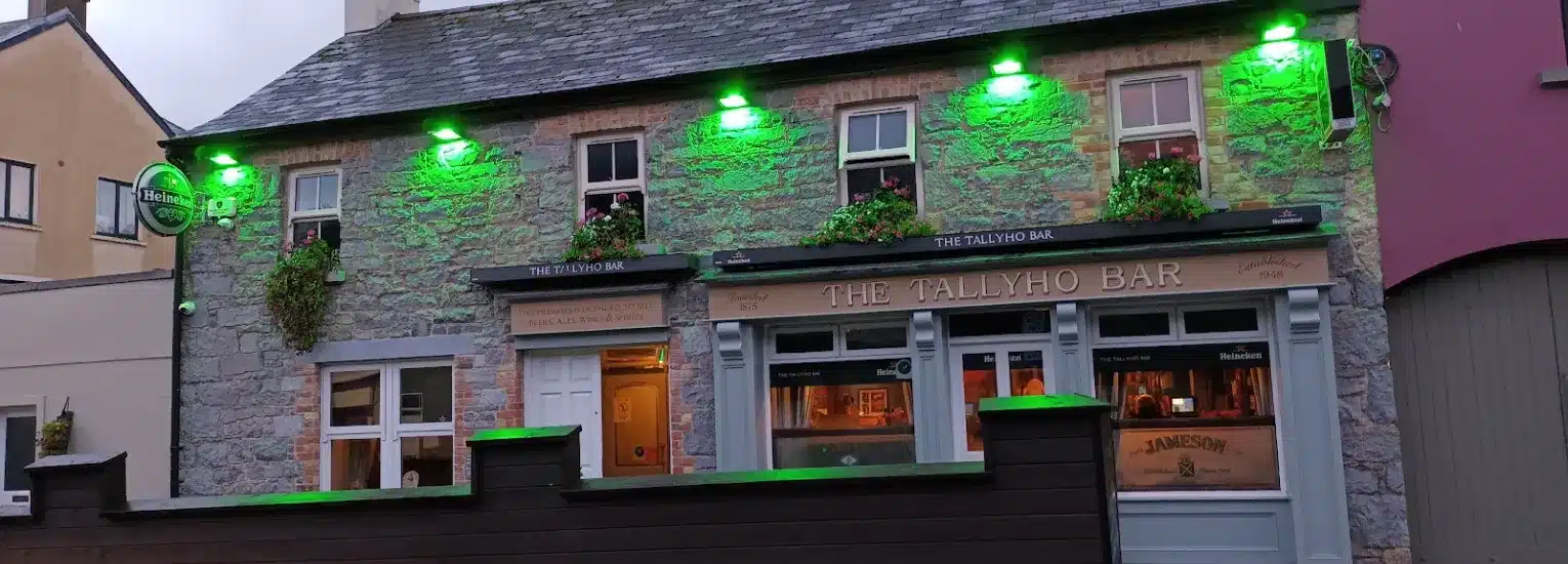 A stone building houses The Tallyho Bar, with green lights illuminating its exterior. The pub has flower boxes on window sills and warm orange lights glowing from inside, creating a cosy, inviting atmosphere.