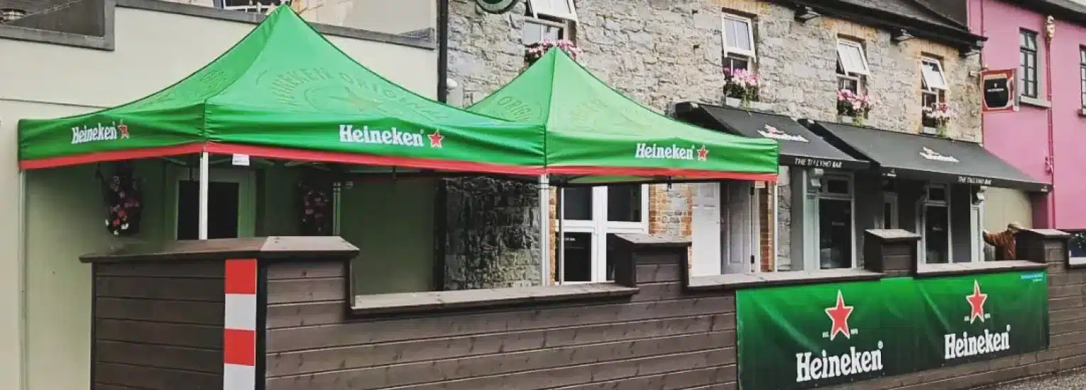 Outdoor seating area of a stone-fronted pub with green Heineken-branded tents and banners, wooden fencing, and flower boxes, located along a street with colourful buildings.