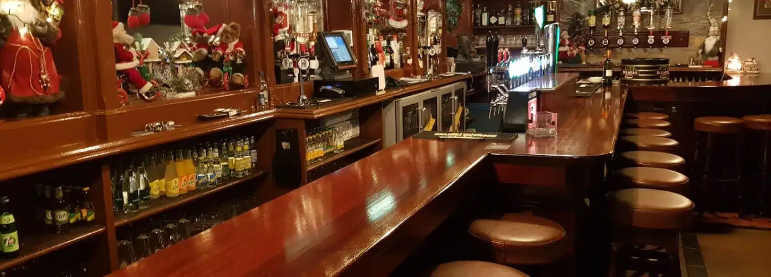 A warmly lit pub with a polished wooden bar, high stools, and shelves stocked with drinks. The space is decorated with festive holiday ornaments and soft toys, creating a cosy atmosphere.