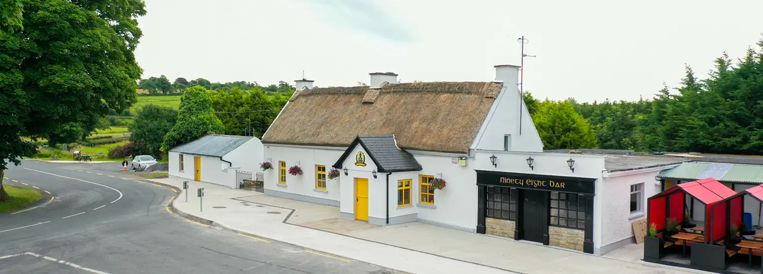 A traditional white thatched-roof pub with yellow doors and window boxes sits beside a quiet rural road, surrounded by greenery and fields under a cloudy sky.