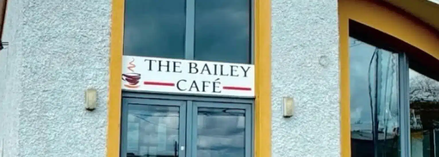 A sign above glass doors reads "The Bailey Café" with a coffee cup logo, mounted on the exterior wall of a modern building with large windows.