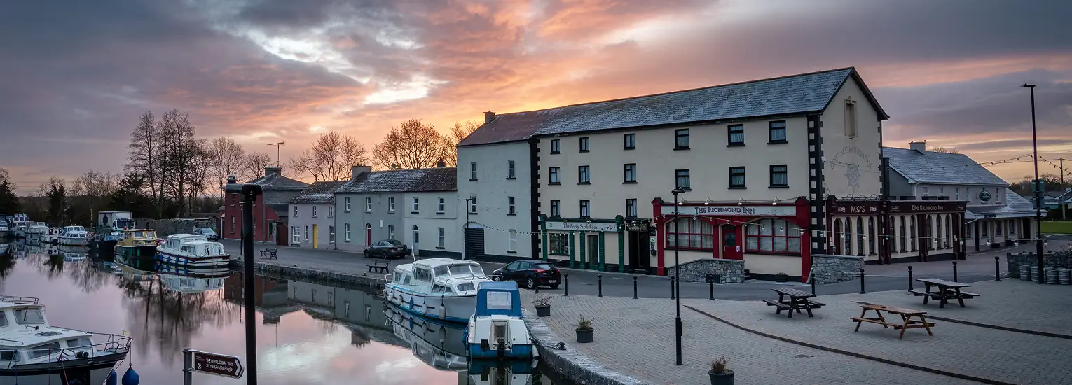 A peaceful riverside scene at sunset shows boats moored along the calm water, with a row of traditional buildings, including restaurants and shops, lining the promenade under a dramatic, colourful sky.