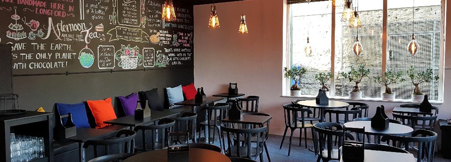 A cosy café with black tables and chairs, colourful cushions on a bench along the wall, chalkboard menus with drawings, pendant lights, and potted plants on the windowsill.