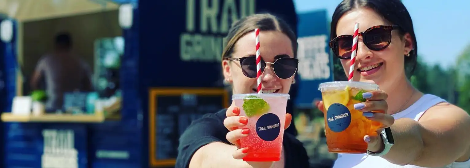 Two women wearing sunglasses smile and hold up colourful drinks with striped straws in front of a food van on a sunny day.