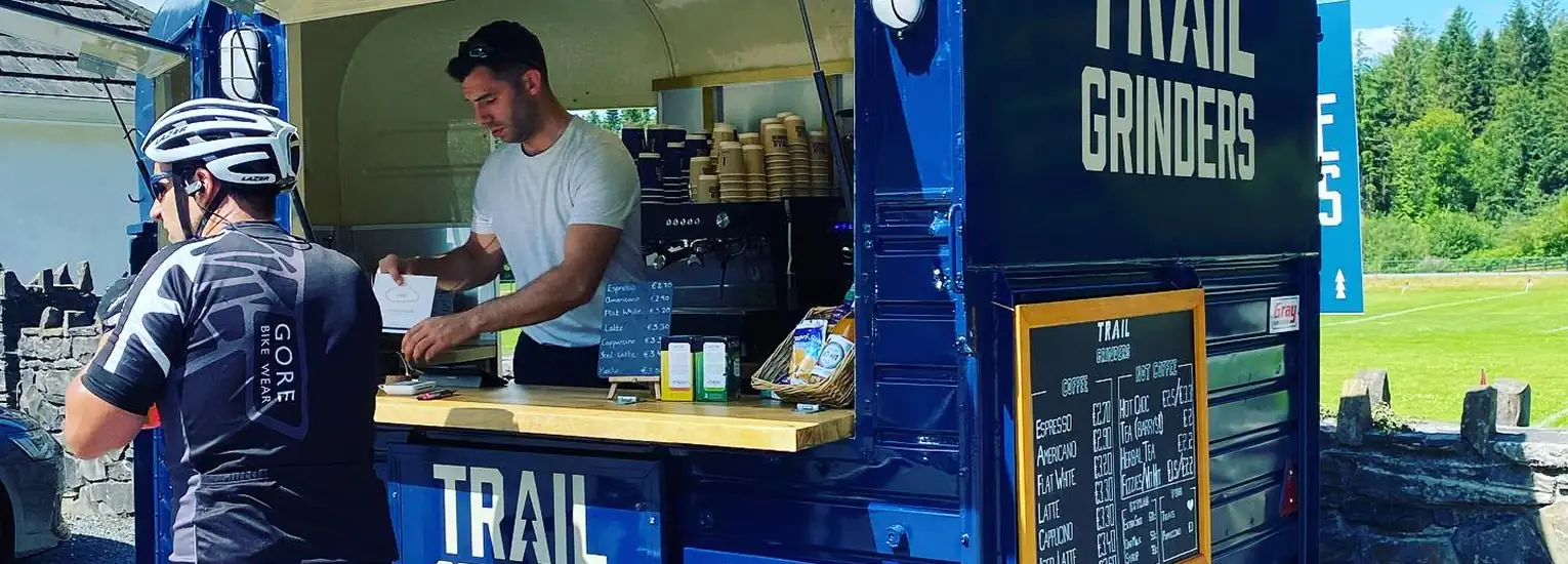 A cyclist orders at a blue coffee van called Trail Grinders, parked outdoors. A staff member prepares a drink inside, with coffee cups and menus visible by the open service window. Trees and grass are in the background.