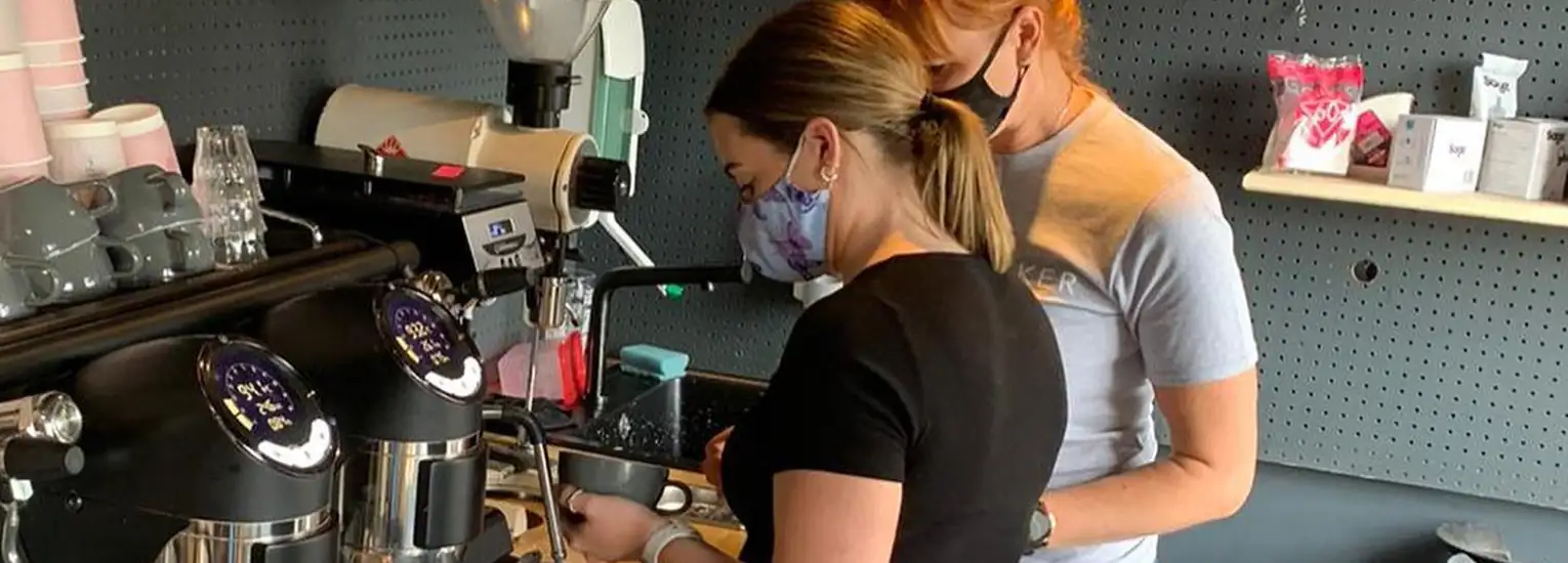 Two women wearing face masks prepare coffee drinks behind a counter in a café. One operates an espresso machine while the other observes. Shelves with cups and supplies are visible in the background.