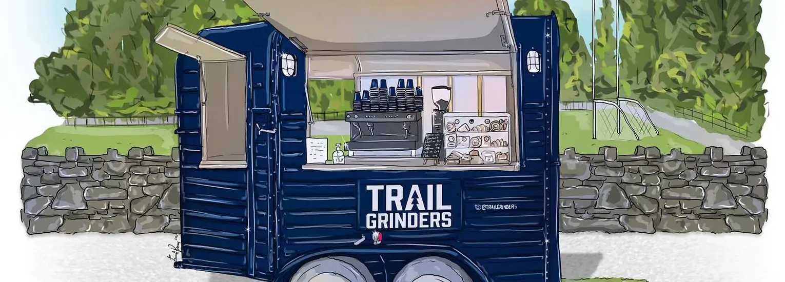 Illustration of a navy blue mobile coffee trailer labelled "Trail Grinders," parked in front of a stone wall, with trees and grass in the background. The trailer window is open, showing a coffee machine and cups inside.