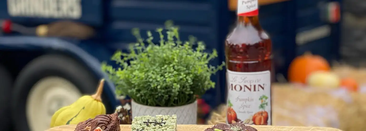 A wooden board with assorted doughnuts sits on an outdoor table next to a potted herb, a bottle of Monin Pumpkin Spice syrup, and autumn decorations, including pumpkins and squash, with a blue vehicle in the background.