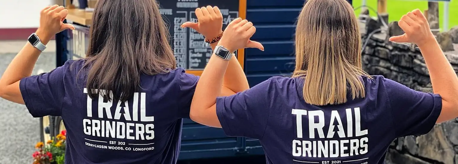 Two people with shoulder-length hair face away from the camera, pointing at the backs of their navy shirts that read "TRAIL GRINDERS." Both wear smartwatches and stand outdoors near a blue container.