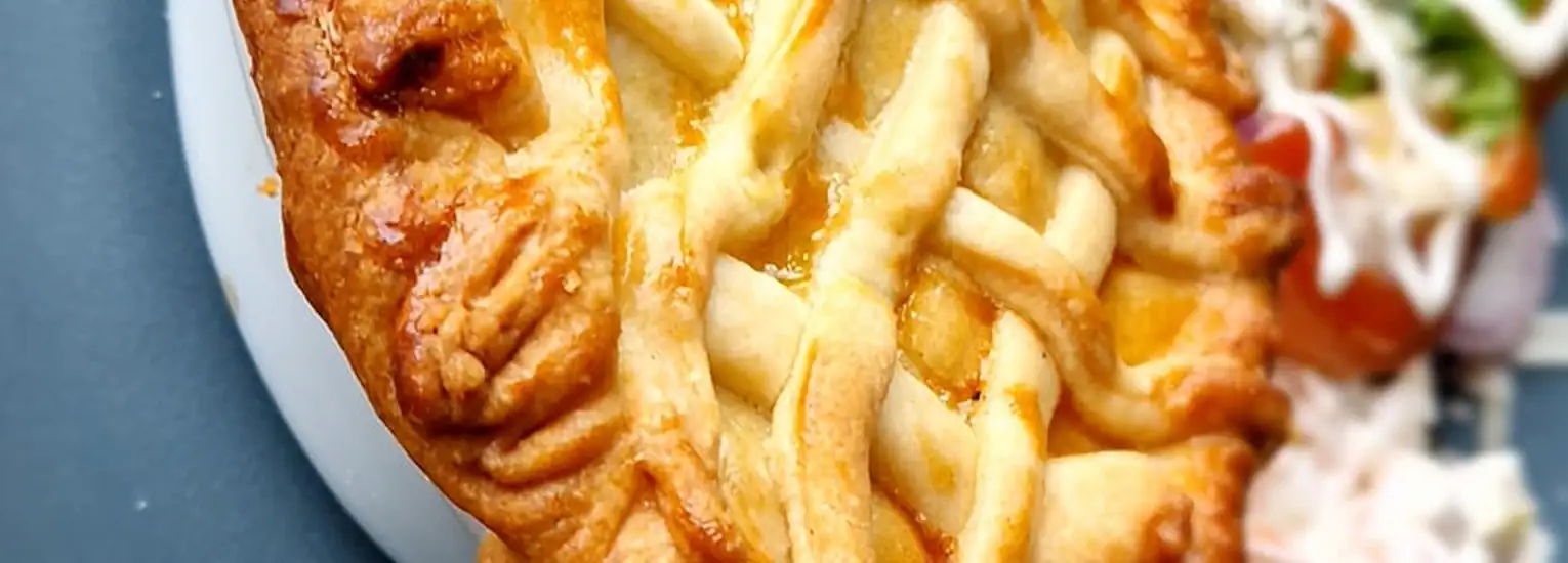 A close-up of a golden-brown lattice-topped pie on a white plate, with a side of salad and cream visible in the blurred background.