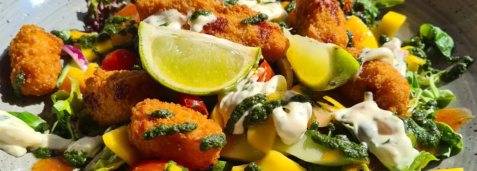 A close-up of a colourful salad with breaded chicken pieces, lime wedges, cherry tomatoes, greens, yellow pepper, and drizzles of creamy and green sauces on a grey plate.