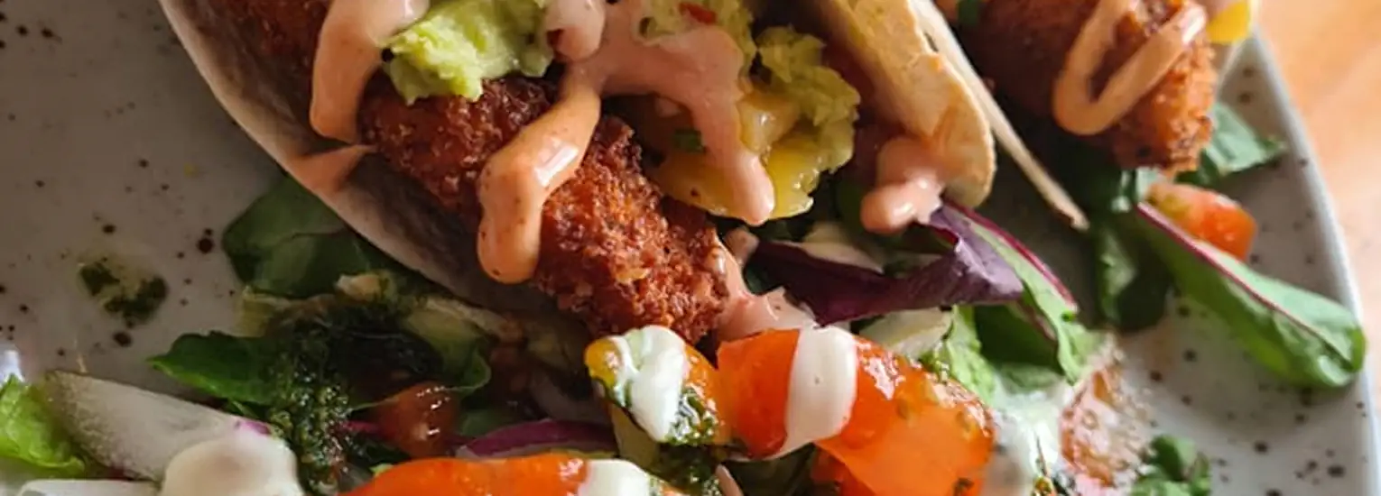 Two tacos filled with crispy breaded pieces, guacamole, and pink sauce, served on a plate with a colourful side salad topped with creamy dressings and fresh greens.