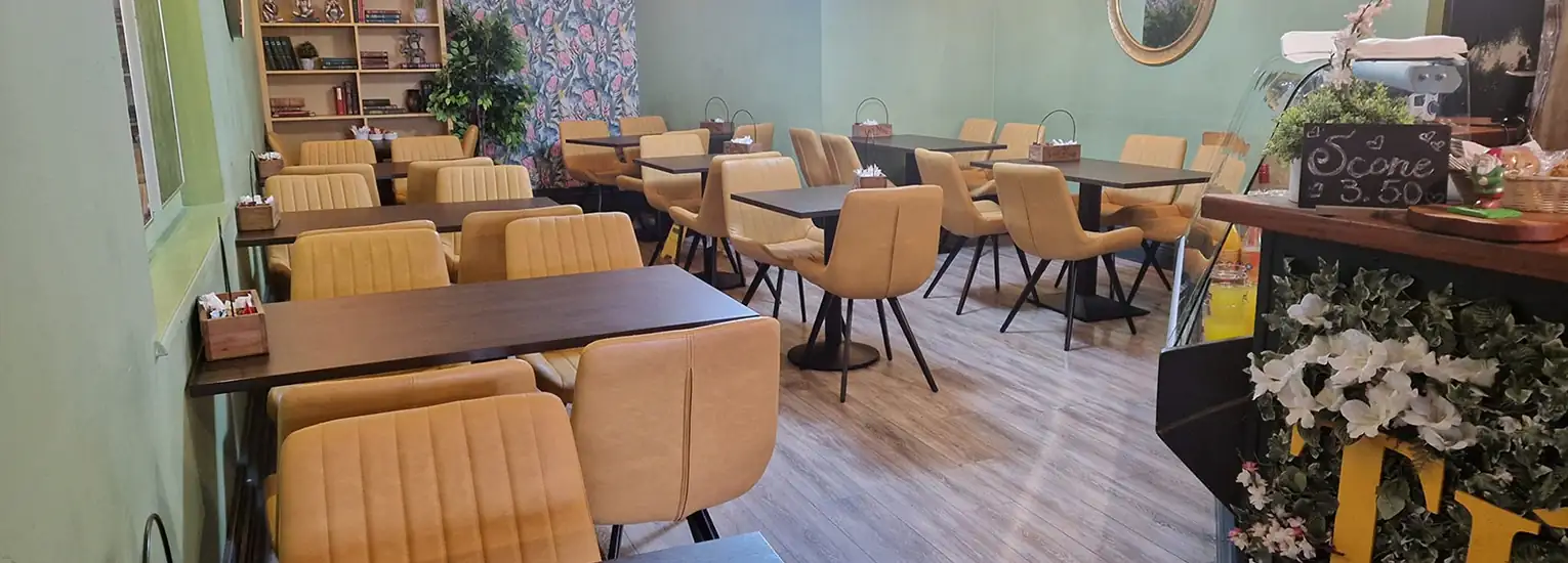 A cosy café with yellow cushioned chairs and dark wooden tables, decorated with plants, bookshelves, a green accent wall, and a floral wallpaper. A counter with flowers and a chalkboard sign is visible in the foreground.