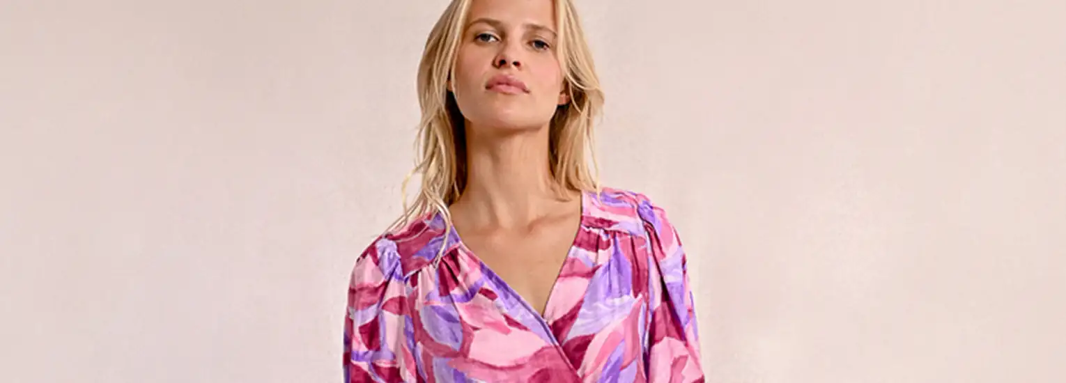 A woman with blonde hair wearing a long-sleeved, pink and purple patterned blouse stands against a plain light background, looking confidently at the camera.