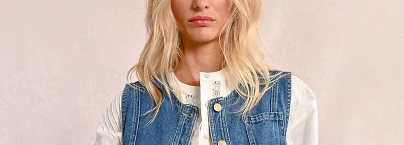 A person with long blonde hair wears a blue denim waistcoat over a white shirt, standing against a light-coloured background.