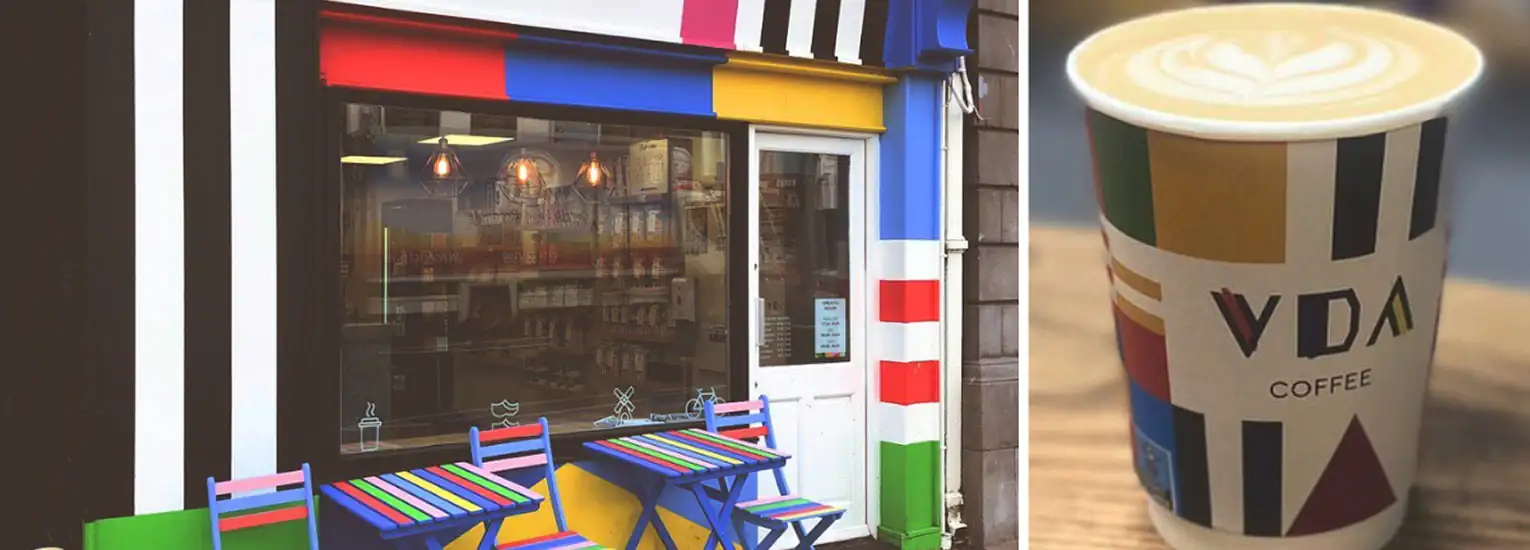 A colourful café exterior with striped designs, two matching tables and chairs outside, and a coffee cup with similar patterns and a latte art heart on top. The cup reads “VDA Coffee.”.