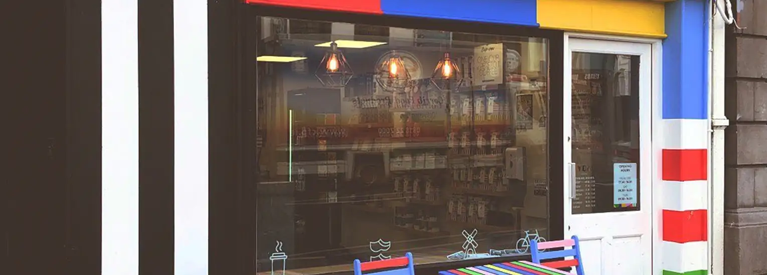 The image shows the exterior of a shop with a colourful striped design in red, blue, yellow, white, and black. There is a large window, three hanging lights inside, and a white door to the right. A striped bench sits outside.