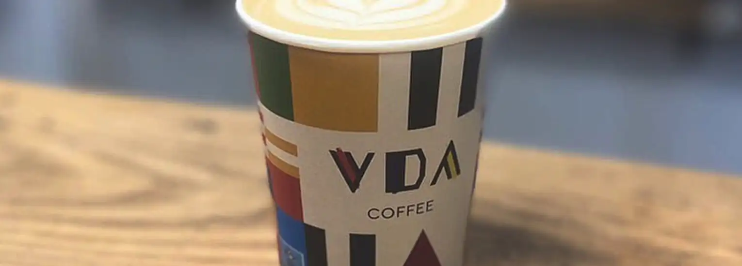 A paper coffee cup with a colourful geometric pattern and "VDA COFFEE" printed on it sits on a wooden surface, filled with coffee topped with latte art.