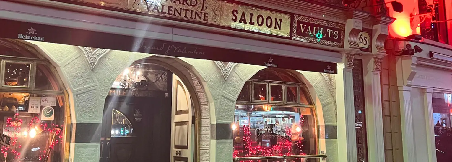 A warmly lit bar with arched windows and a sign reading "Valentine Saloon" at night. Red fairy lights and heart-shaped decorations are visible inside, creating a cosy, festive atmosphere.