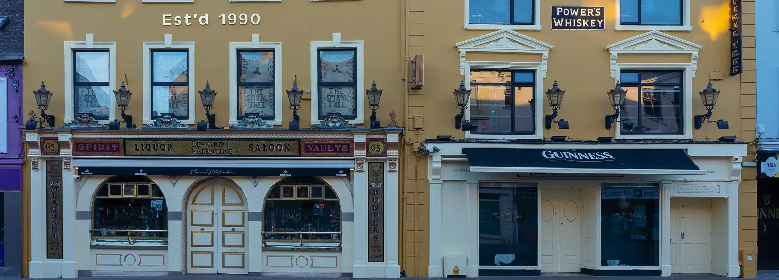 Two adjacent pubs with yellow façades, vintage lamps, and signage for Guinness and Powers Whisky. The left pub has arched windows and doors, with "Est'd 1990" above. Both buildings have ornate details and closed windows.