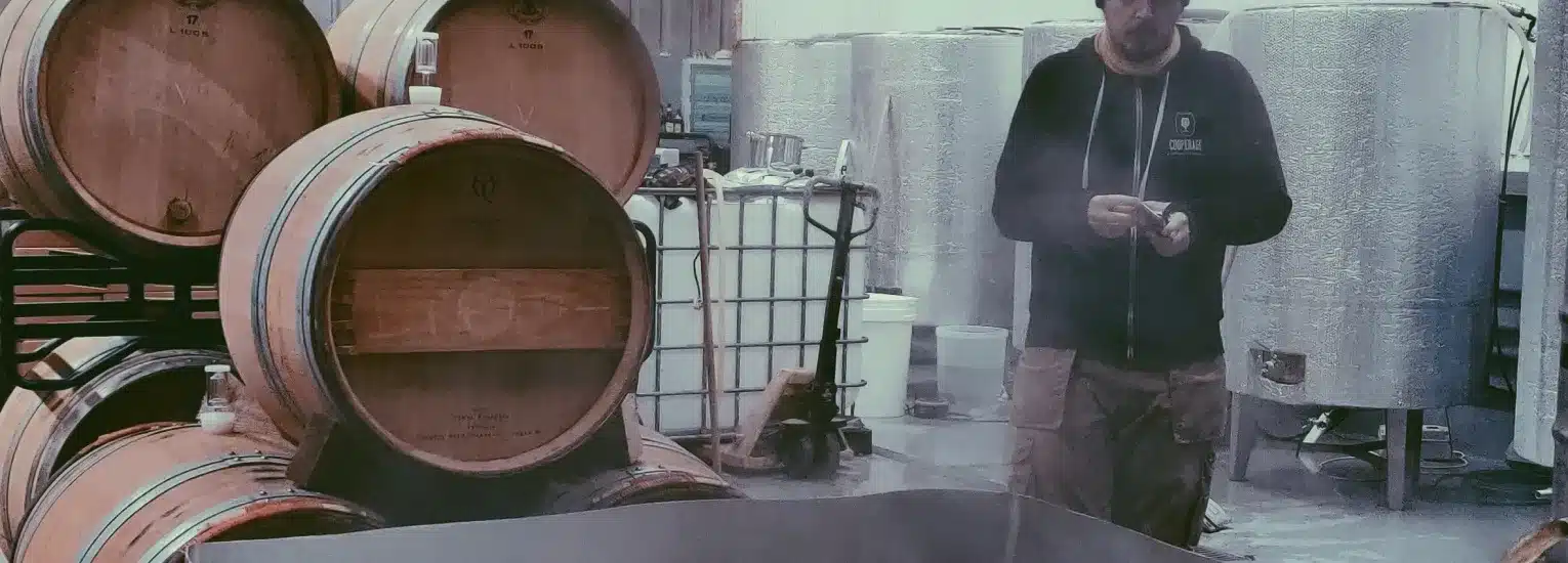 A person stands in a winery or brewery, surrounded by wooden barrels and large metal tanks, with steam rising in the foreground, suggesting a process involving heat or fermentation.