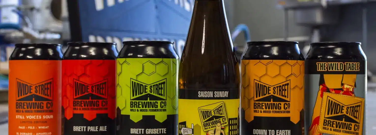 Five colourful cans and one brown bottle of Wide Street Brewing Co. craft beer are lined up on a table. The cans and bottle display various beer names and vibrant labels with the brewery’s logo.
