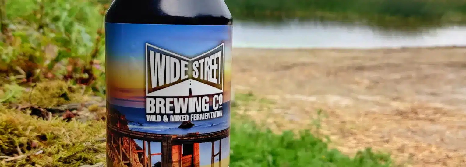 A bottle of Wide Street Brewing Co. beer stands on grass near a calm lakeshore, with trees and water in the background. The label features a scenic image and bold text.