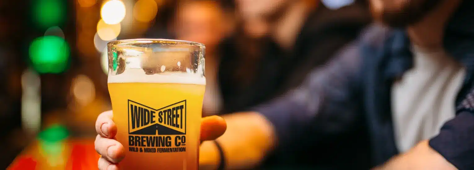 A person holds a pint glass of hazy beer with a "Wide Street Brewing Co." logo at a bar; the background is softly blurred with warm lighting and other people present.