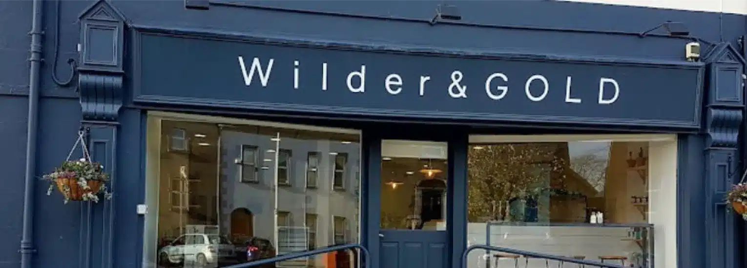 Shopfront with a dark blue sign reading "Wilder & GOLD" in white letters, large glass windows showing reflections of buildings and some interior details, and hanging flower baskets on either side.