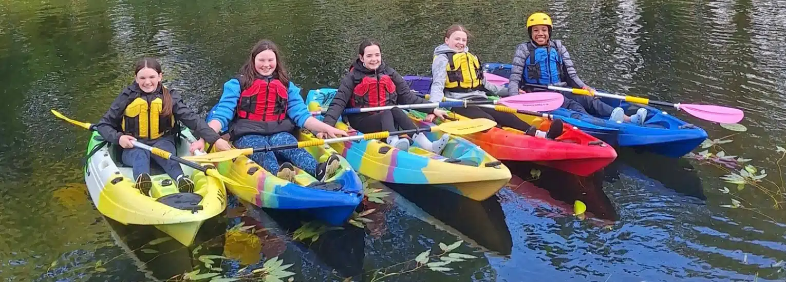 Five people in colourful kayaks, wearing life jackets and holding paddles, smile on a calm body of water. One person wears a yellow helmet. Green leaves float nearby.
