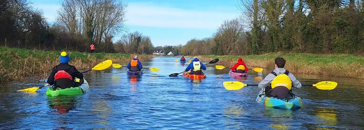 A group of people in colourful kayaks paddle down a calm river lined with grass and trees under a blue sky.
