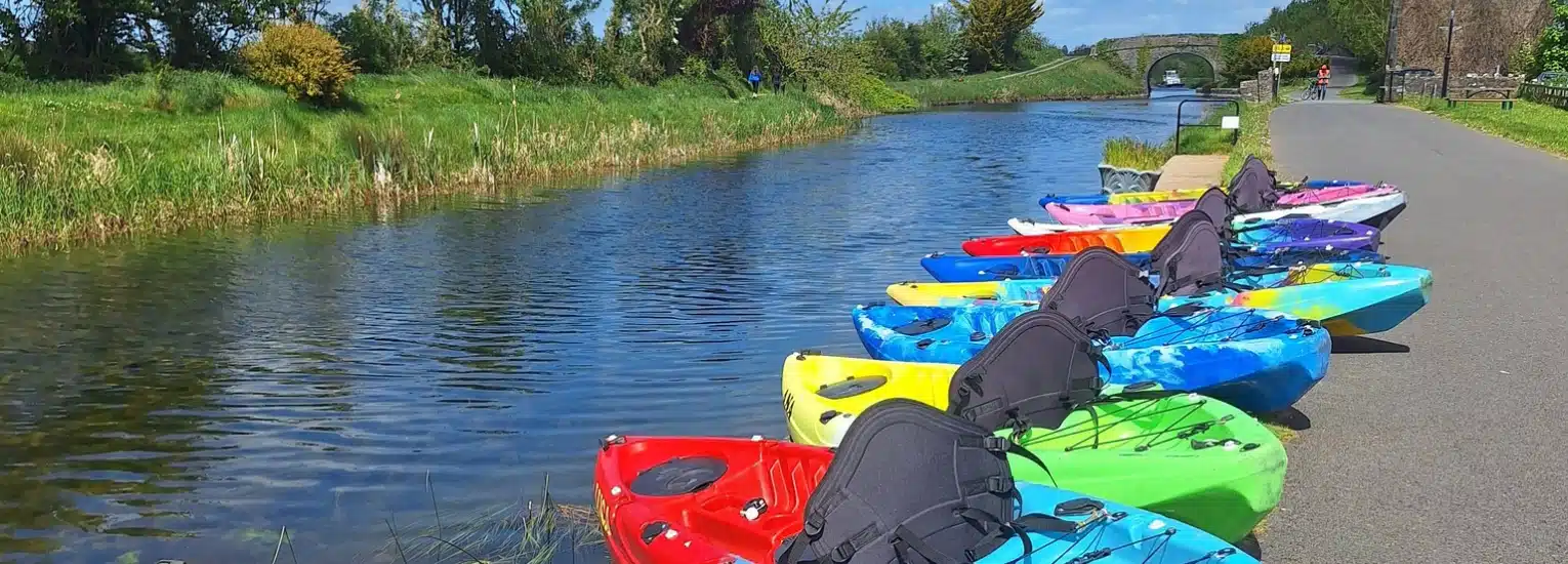 Colourful kayaks lined up along the edge of a calm canal, with a paved path and greenery on one side, and a stone bridge visible in the distance under a blue sky.