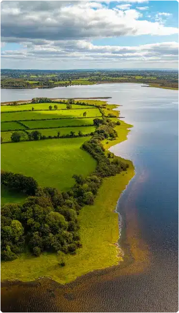 Aerial view of lush green fields bordered by hedges and trees beside a large, calm river or lake under a partly cloudy sky.