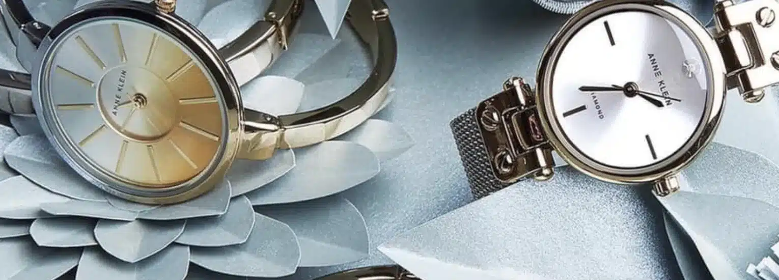 Two Anne Klein wristwatches placed on silver paper flowers; one watch has a gold face with gold strap, the other has a white face with gold mesh strap, both reflecting soft light on a light blue background.
