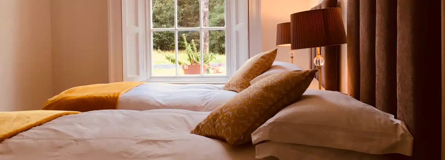 A cosy bedroom with two single beds, each adorned with white linen, yellow throws, and patterned cushions. A window between the beds lets in natural light, and a lamp stands in the corner beside brown curtains.