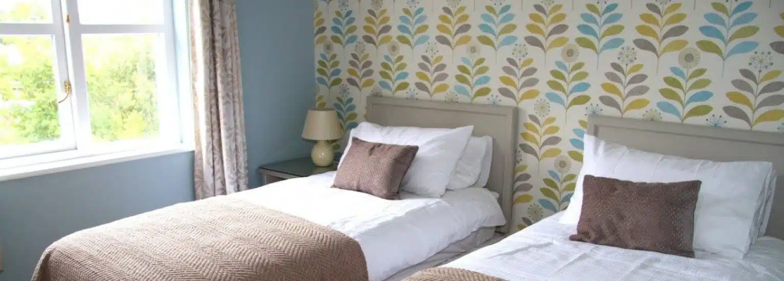 A bright bedroom with two single beds, each with white bedding, beige throws, and brown cushions. A lamp sits on a bedside table, and a window with curtains lets in natural light. The wallpaper features a colourful leaf pattern.