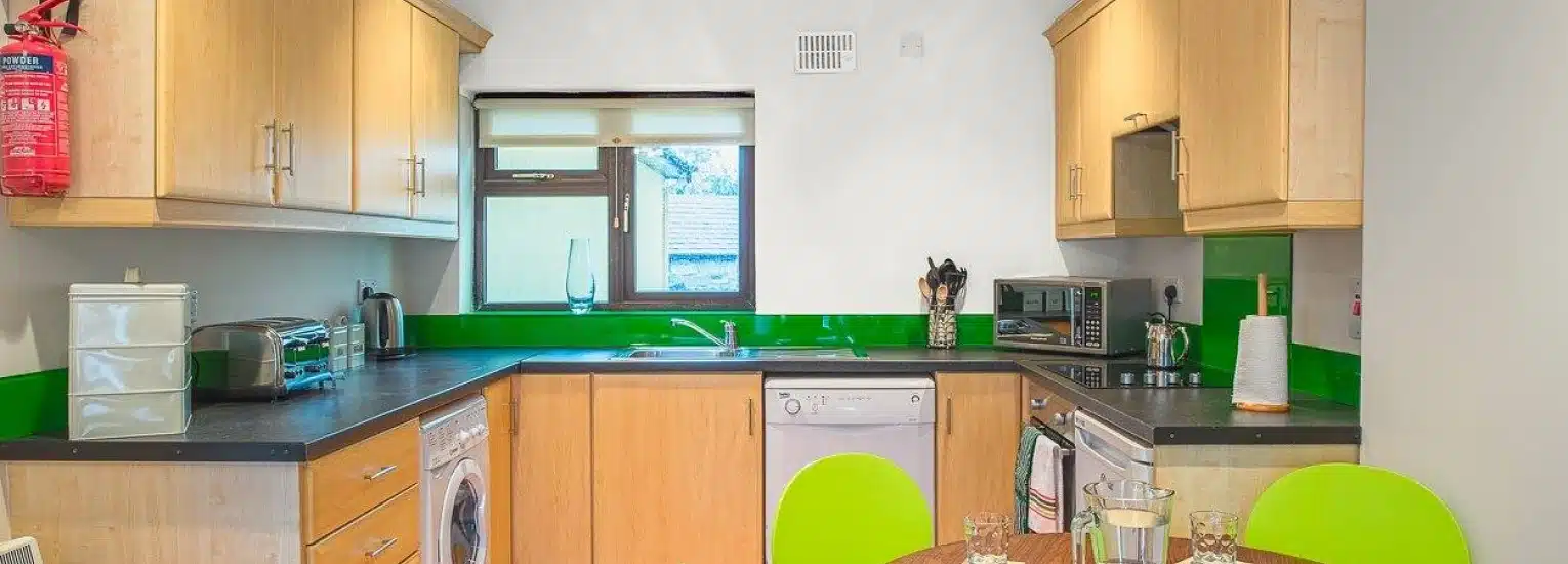 A modern kitchen with light wood cupboards, green accents, a worktop with appliances, a small table with two chairs, and a window above the sink letting in natural light.