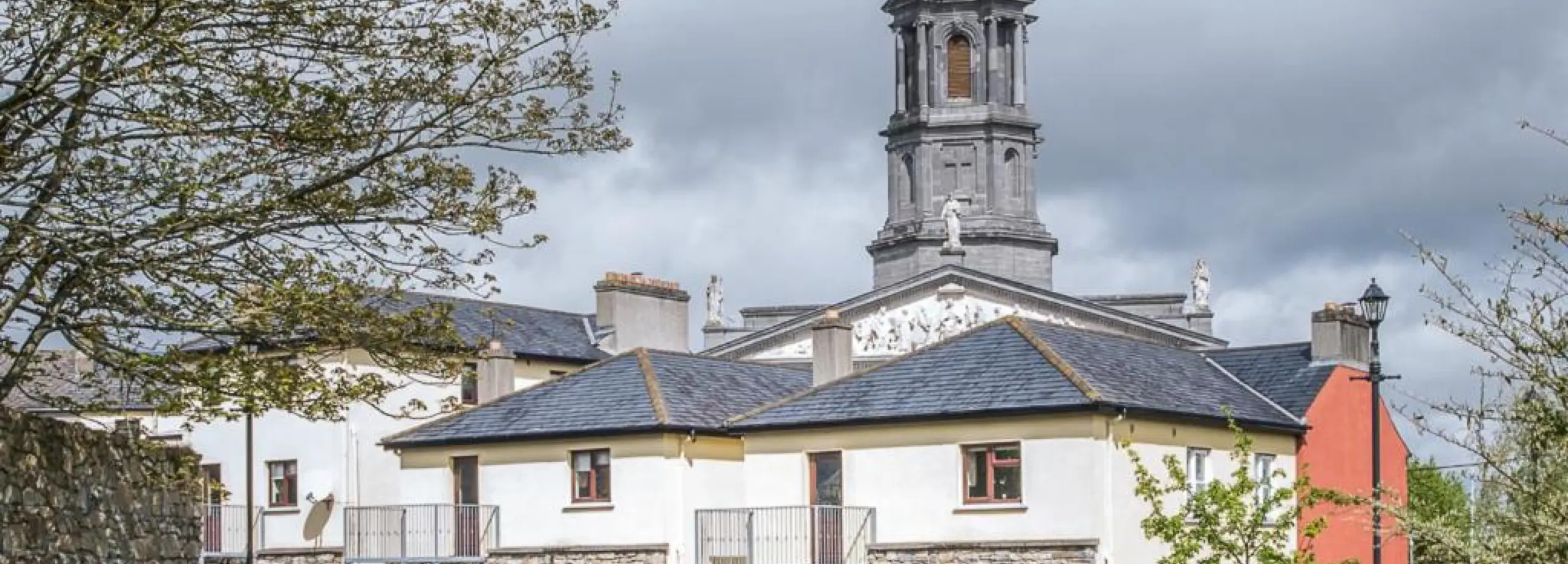 A stone church tower rises behind cream-coloured buildings with slate roofs, framed by leafy branches and a cloudy sky in the background.