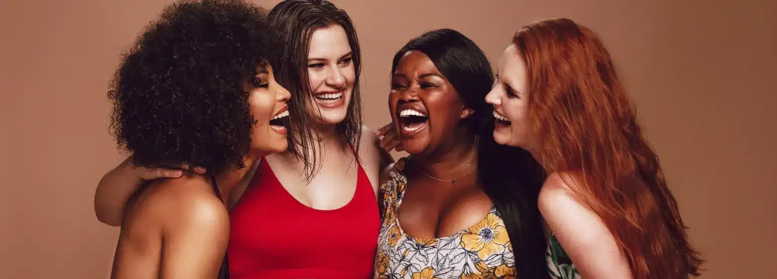 Four women with diverse skin tones and hair types are standing close together, smiling and laughing against a neutral background, showing joy and friendship.
