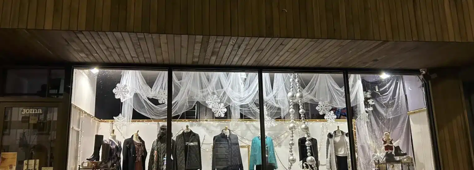 A clothing shop window display at night features mannequins dressed in winter clothing. The backdrop is decorated with white sheer fabric and large snowflakes, creating a festive, wintry atmosphere.