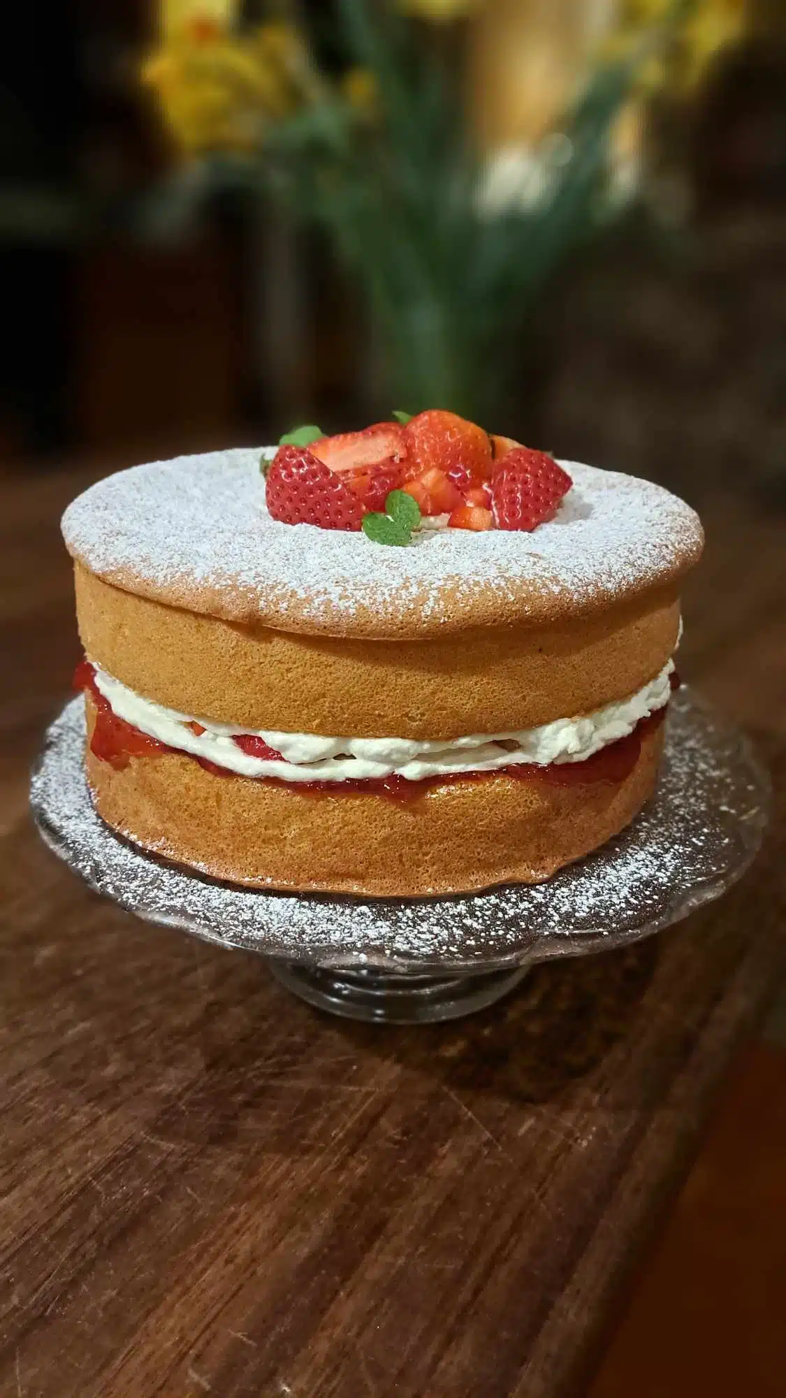 A Victoria sponge cake sits on a glass stand. It has a layer of cream and jam in the middle, is dusted with icing sugar on top, and is decorated with sliced strawberries and small green leaves.