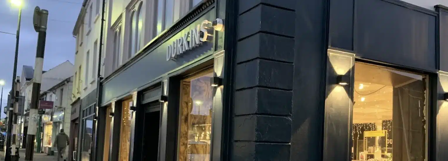 A dark-painted shopfront with large windows illuminated from inside, displaying various items. The street is lined with other buildings, lamp posts, and a few pedestrians in the evening light.