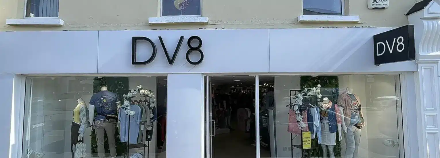 Street view of a clothing shop called DV8, with large glass windows displaying mannequins dressed in casual outfits and jackets, and the shop entrance in the centre. The shop's name appears above the entrance and on a side sign.