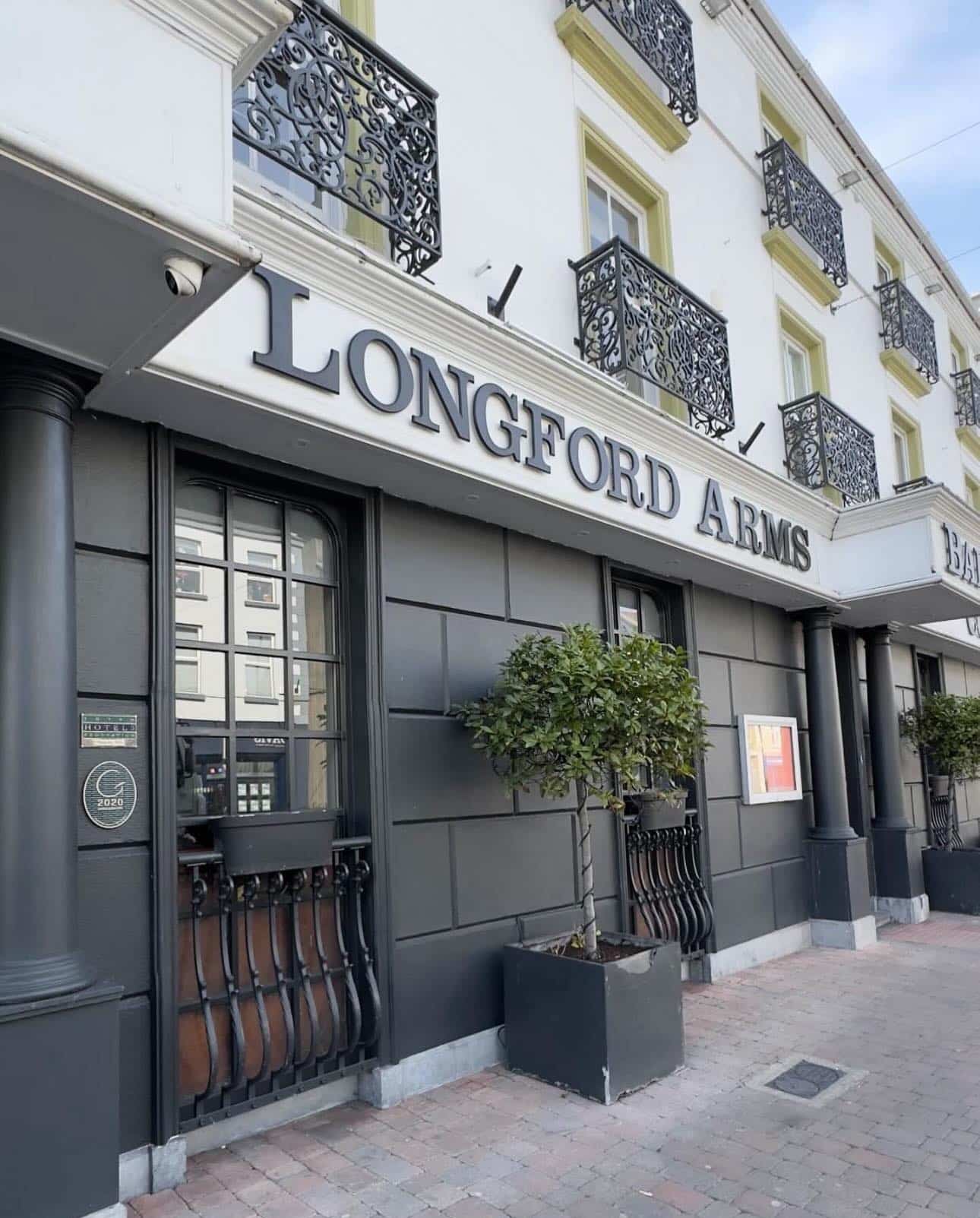 A hotel or pub exterior with a sign reading "LONGFORD ARMS," featuring decorative black railings, potted trees, and ornate balcony railings on the upper storeys.